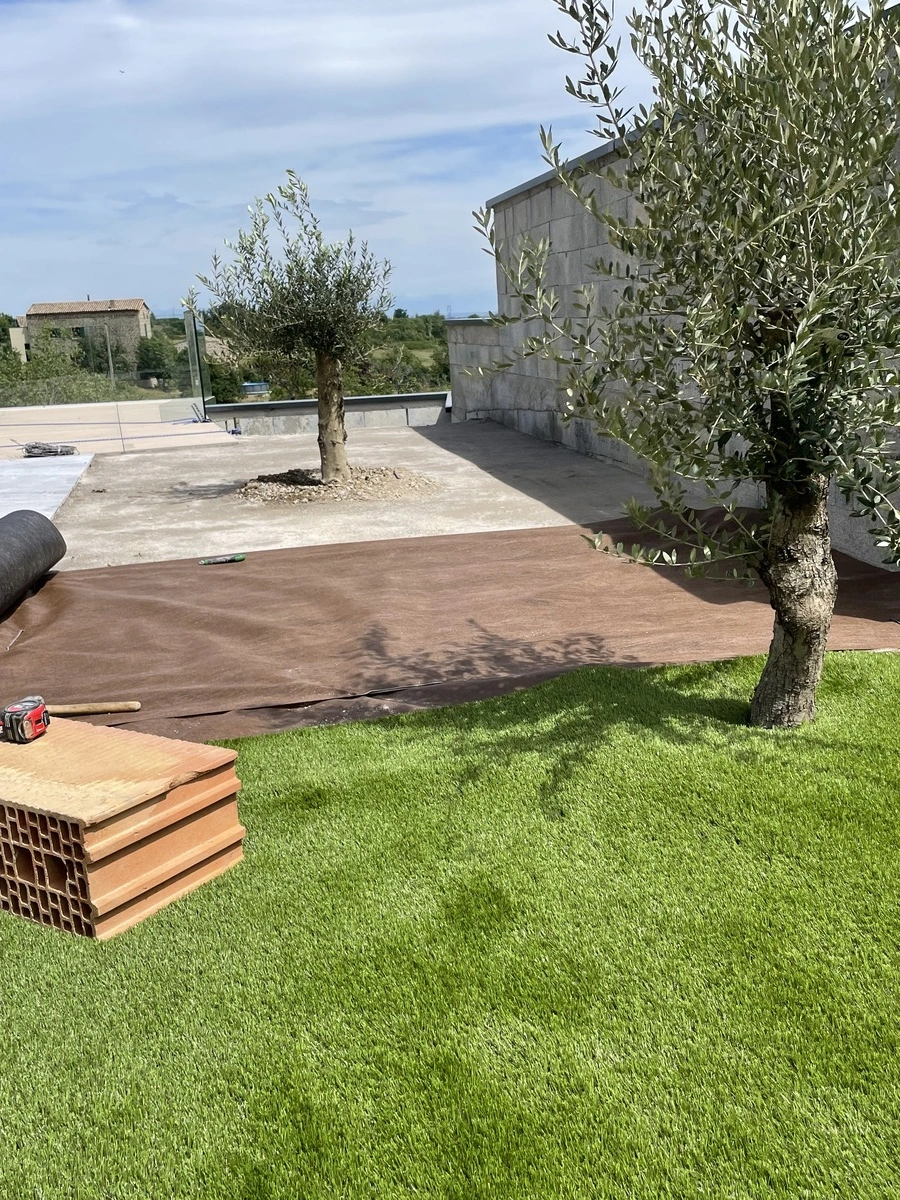 Zone de jardin en cours d'aménagement avec une pelouse verdoyante, des oliviers et des matériaux de construction, montrant les étapes d'un projet d'aménagement paysager pour espaces verts.